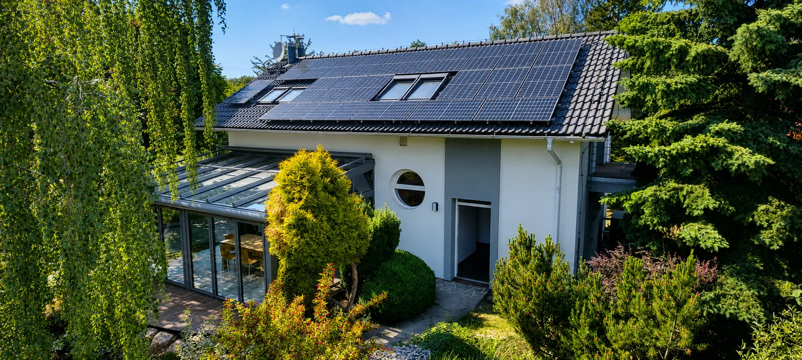 Exteriér domu Včelník 322 – bílá fasáda s prosklenou zimní zahradou Solarlux, terasa a fotovoltaika na střeše, obklopeno vzrostlou zelení.